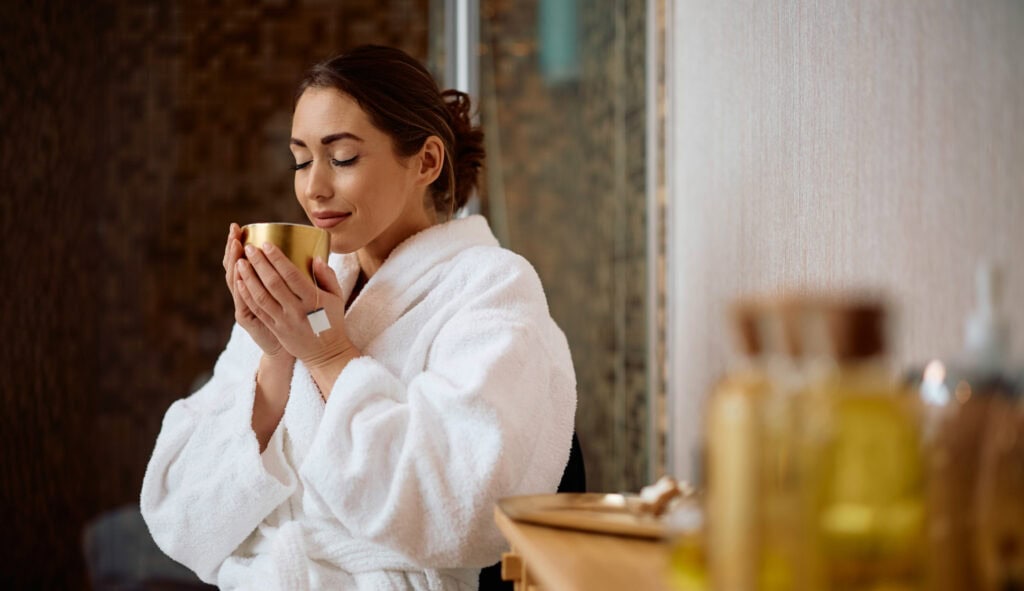 Relaxed woman in a white bathrobe enjoying a cup of tea or coffee in a cozy setting.